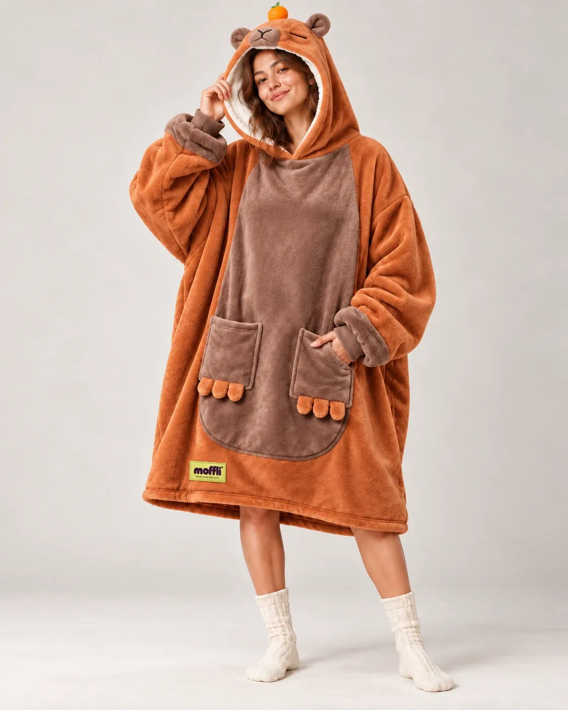 Woman standing wearing the orange and brown Capybara Moffli, hood up with face, ears and mandarin, paw pockets and Moffli label